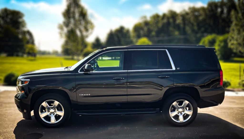 The Private Tahoe vehicle is a Chevy Tahoe, shiny black with silver hubcaps and trim. It is parked in this photo in front of a green field and there are green trees. The sky is blue with fluffy white clouds.