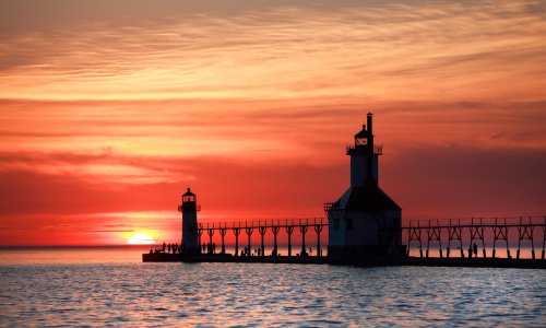 The St. Joseph lighthouse and pier at sunset. The sky is deep orange and the sun is halfway below the horizon over Lake Michigan.
