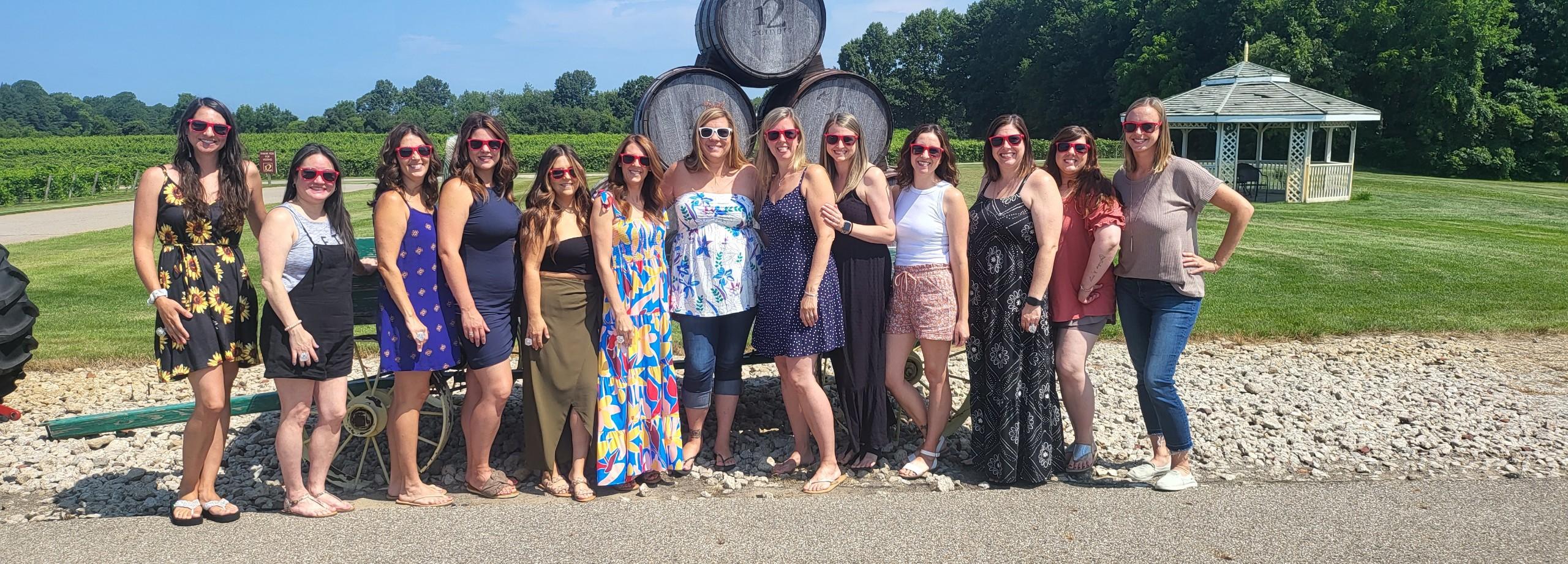 A group of women in summer sundresses stand in front of a group of wine barrels outside at a winery. It is summer, the sky is bright blue, there are green trees and a grassy lawn behind them. There is also a wooden gazebo on the lawn.