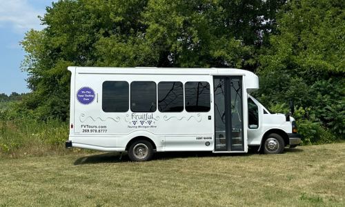 The Vino Coach is a white vehicle with the Fruitful Vine Tours logo on the side. In this photo it is parked in a green field in front of a stand of green trees and a blue sky.