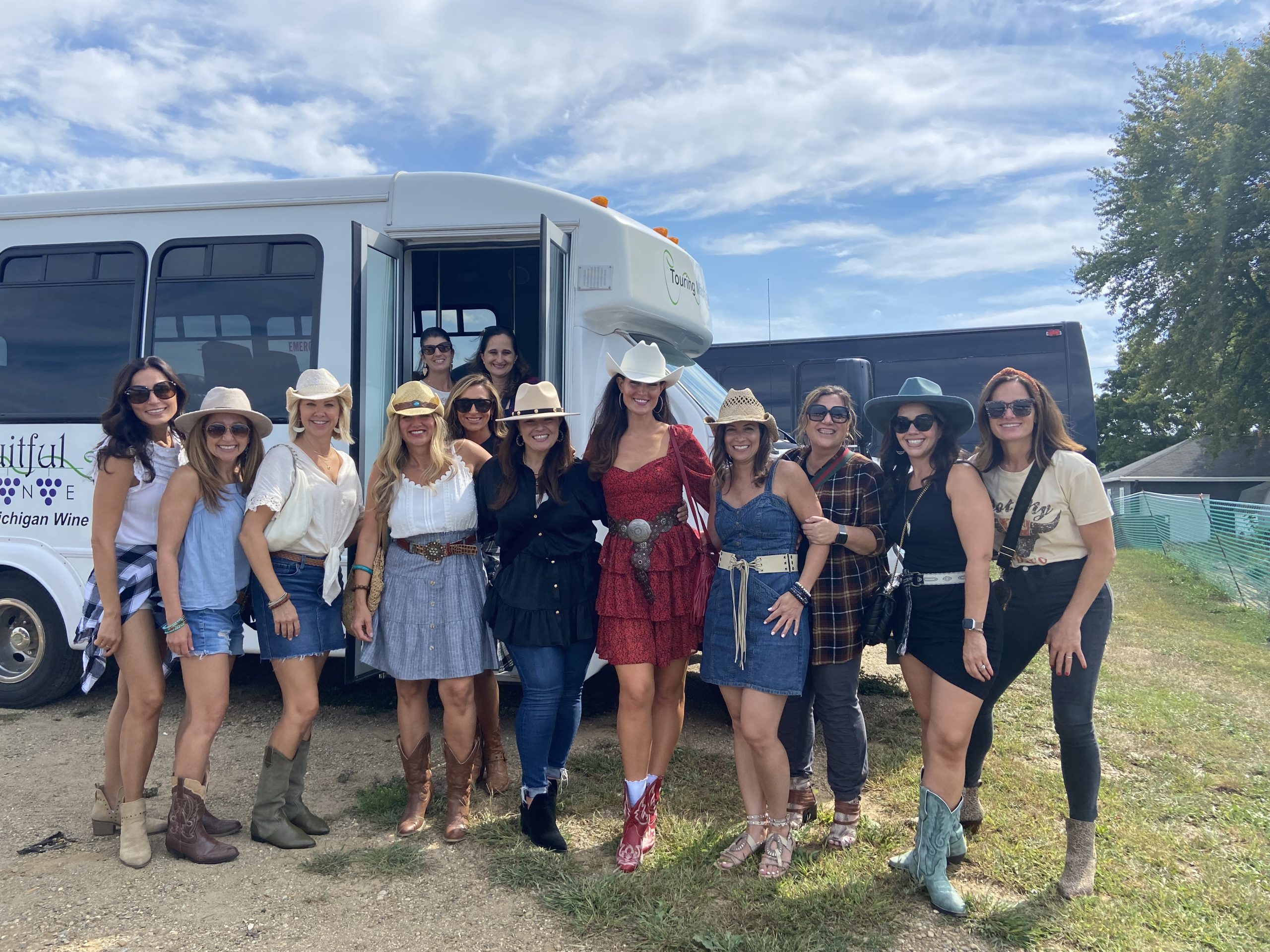 A group of women pose in front of the Fruitful Vine Vino Coach at a winery. It is summer and the women are dressed in country western styles with boots and cowboy hats. The sky is bright blue with clouds, and there is a green tree in the background.