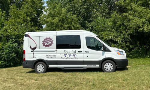 The Private Transit vehicle is a white van with the Fruitful Vine logo on the side. In this photo it is parked in a green field in front of green trees.