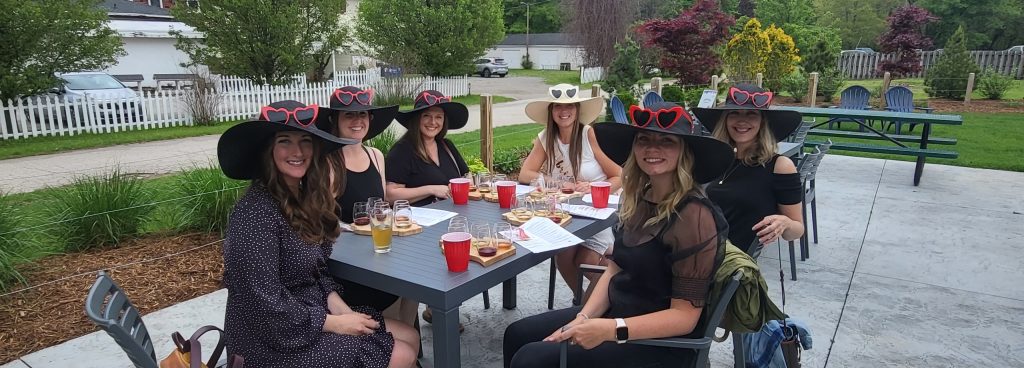 A group of women in fancy dresses and hats enjoy wine at a winery. One woman is wearing white and all the others are wearing black. They also have matching heart-shaped sunglasses. They are sitting outside at a picnic table. There is a green lawn behind them, and some white buildings.
