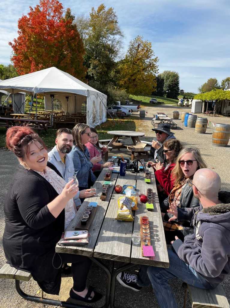 A group of friends enjoy a picnic and wine at a winery. The trees behind their picnic table are varying shade of red, gold, and green and it is early fall. There is also a white tent behind the picnic table.