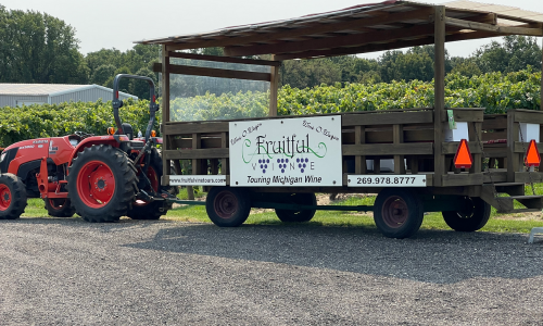 The Wine-O-Wagon. It is an open-air wooden wagon with a roof, pulled by a red tractor. In this photo it is parked in front of a vineyard.